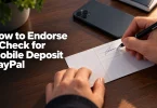 Hands signing the endorsement strip on the back of a check, with the heading How to Endorse a Check for Mobile Deposit PayPal overlaid