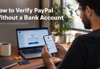 Young man at a modern home office desk holding a smartphone displaying a PayPal-style account verification screen, with a laptop showing an account verification page beside him. Desk holds a notebook, pen, debit card, wireless earbuds, and coffee cup. Soft natural light, clean minimalist setting. Overlay text: “How to Verify PayPal Without a Bank Account – Complete Verification Guide”.